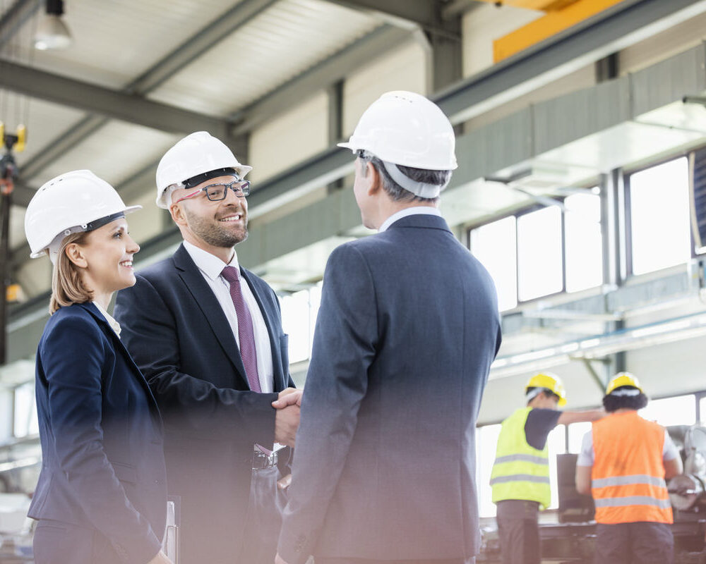 Businessmen,Shaking,Hands,With,Workers,Working,In,Background,At,Metal